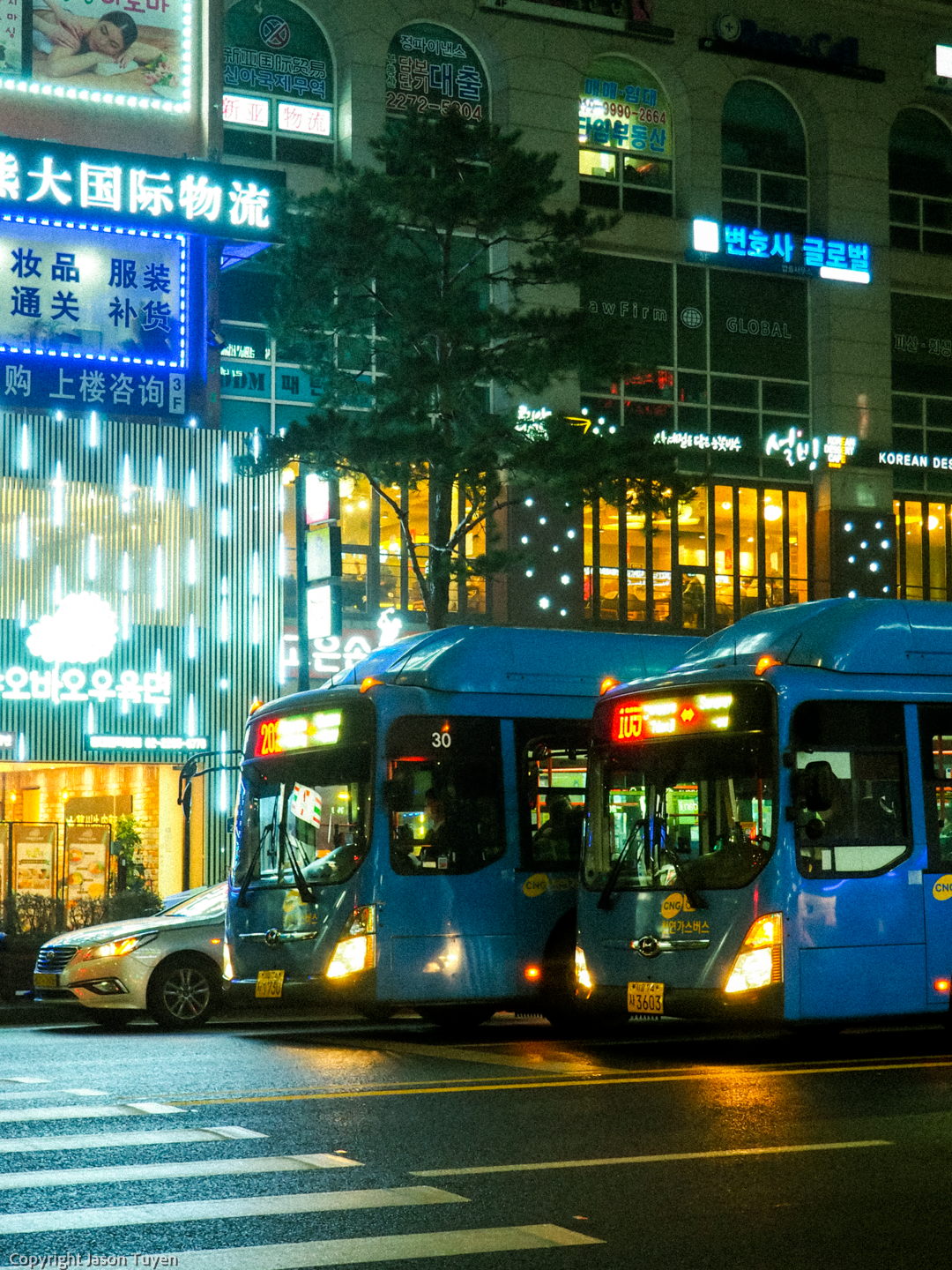Two blue buses stopped at an intersection at night in Seoul.